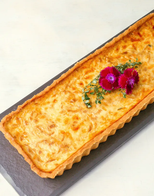 Quiche retangular de cebola caramelizada e queijos, dourada, com flores comestíveis